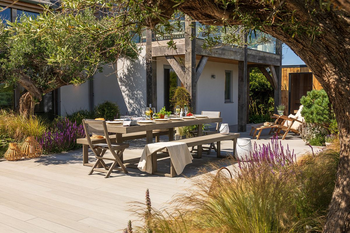 A large outdoor dining table is set for a meal on decking surrounded by plants and trees.