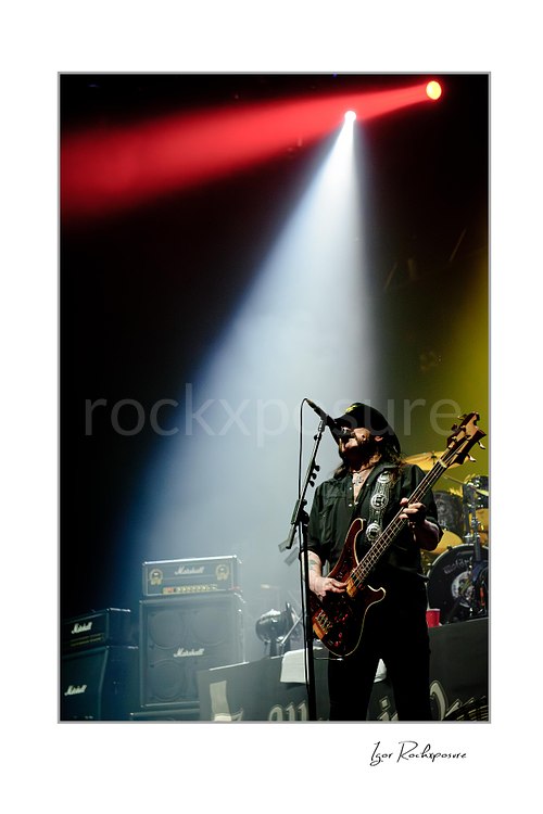 Vertical color image of Lemmy Kilmister of Motörhead performing live on stage with a bass guitar, microphone stand, Marshall amps, and a bright white spotlight