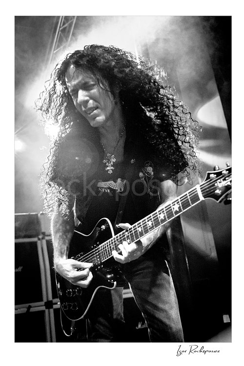 Vertical black and white image of Marty Friedman playing guitar onstage with long curly hair and bright stage haze