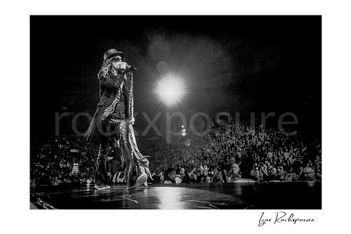 Horizontal black and white image of Steven Tyler of Aerosmith performing live on stage with a scarf-wrapped microphone stand, bright backlight, and a large arena crowd