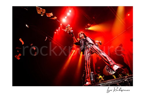 Horizontal color image of Alice Cooper performing live on stage under red lighting with dramatic low-angle perspective and flying stage props