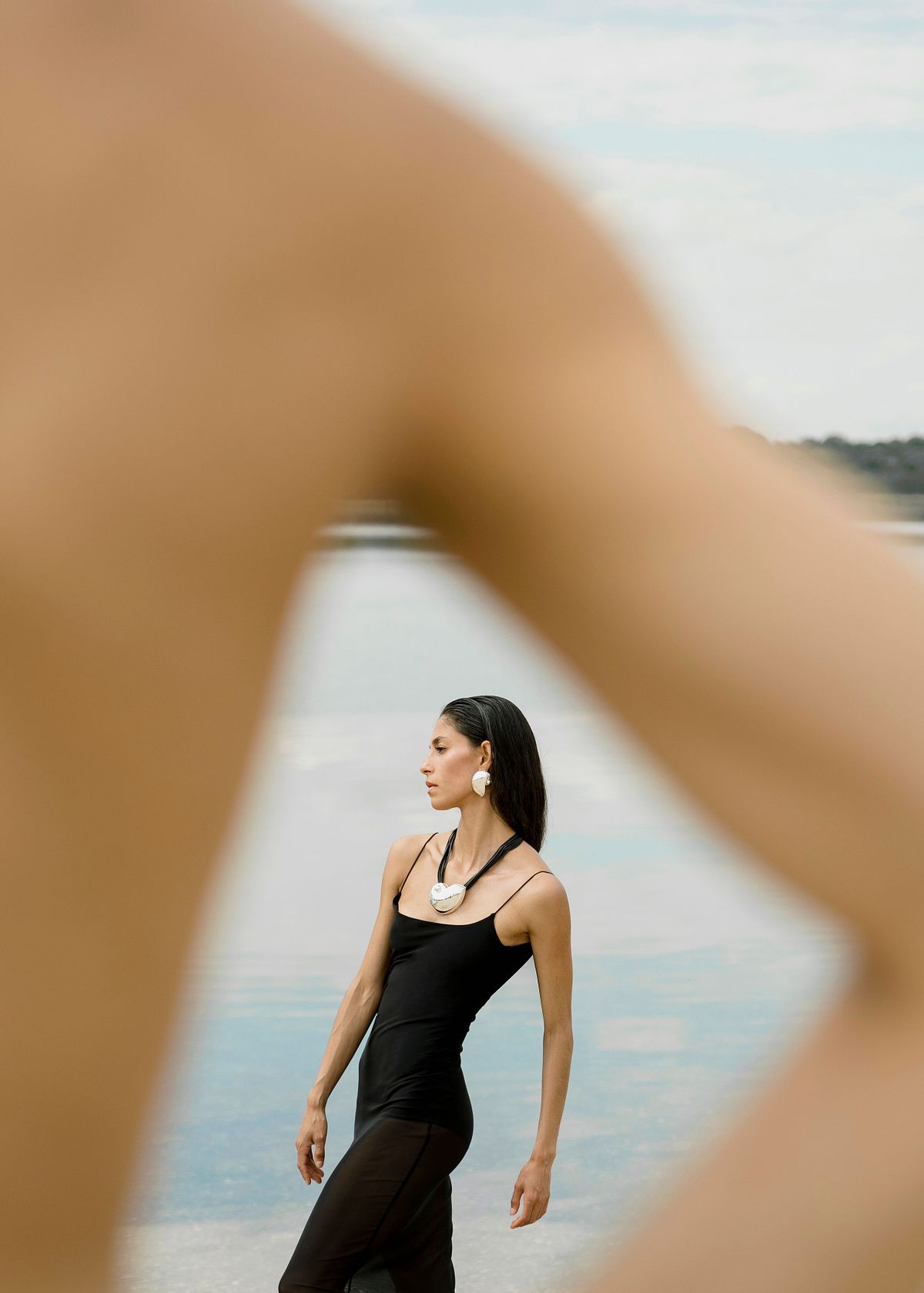 Portrait of a female model with elegance wearing statement jewelry and a black see through summer dress with straps near a lake. Photographed by Filianna Karamanli.