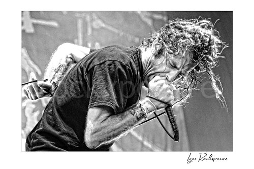 Horizontal black and white image of Randy Blythe of Lamb of God in a tight stage close-up screaming into a handheld microphone with wet hair