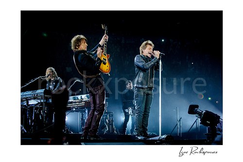 Horizontal color image of David Bryan, Richie Sambora, and Jon Bon Jovi performing live on stage, with Sambora playing guitar and Jon singing at the microphone under cool blue arena light