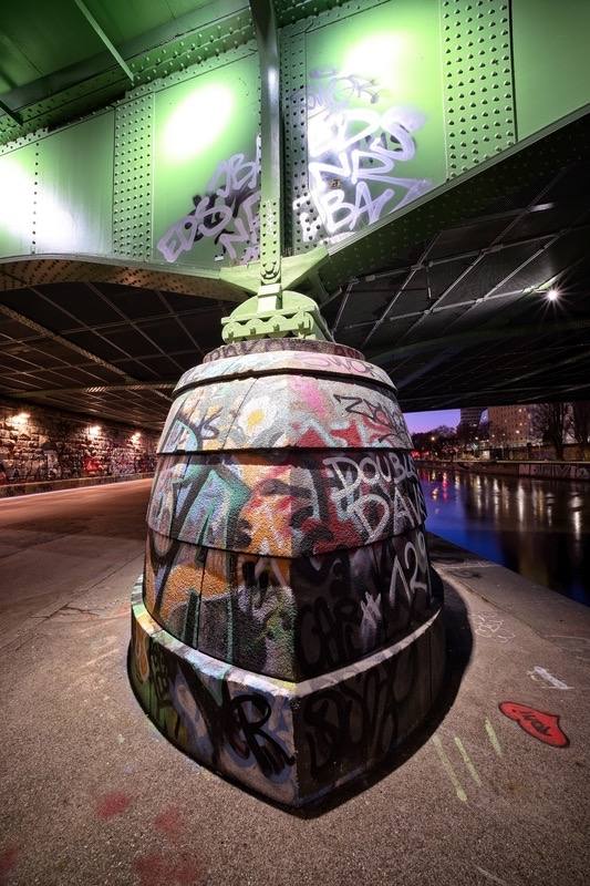 Urbaner Fine-Art-Print aus Wien: Buntes Graffiti auf einem massiven Brückenpfeiler unter der grünen Stahlkonstruktion am Donaukanal bei Nacht