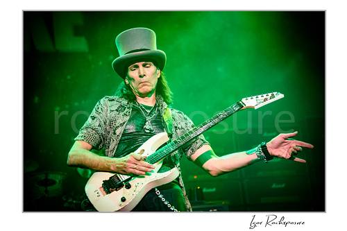 Horizontal color image of Steve Vai playing guitar in a tall top hat under vivid green stage lighting with an outstretched hand gesture