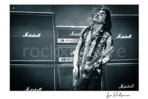 Horizontal black and white image of Nuno Bettencourt performing live on stage with an electric guitar, windblown hair, and a wall of Marshall amplifiers behind him