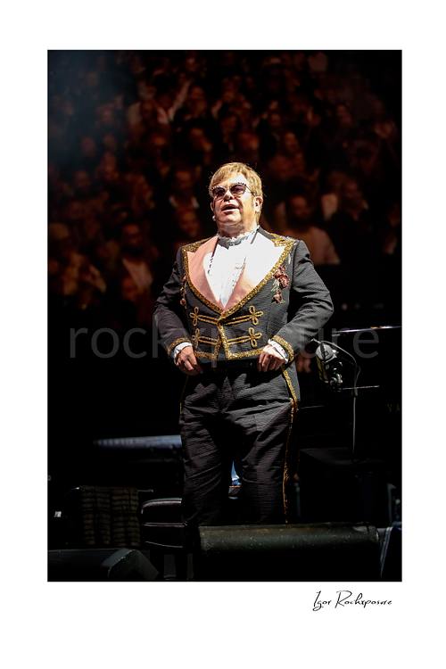 Vertical color image of Elton John standing on stage before a blurred concert crowd in an ornate performance suit