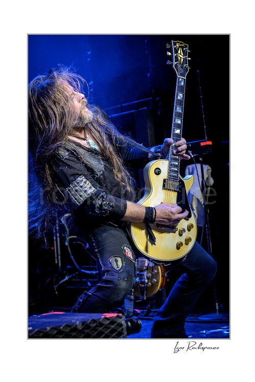 Vertical color image of Chris Latham of Beasto Blanco playing guitar under vivid blue stage light, with long hair in motion and a cream Les Paul style guitar