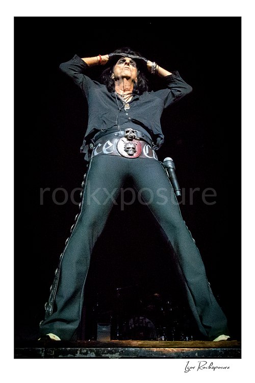 Vertical color image of Alice Cooper performing live on stage in a black outfit with hands raised above his head and a large skull belt buckle