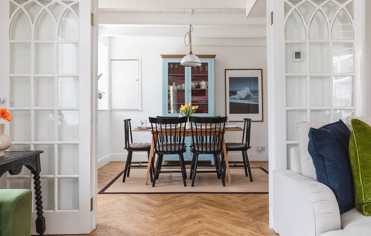 Dining room with a wooden table, six chairs, a pendant light above, and a vase of yellow tulips, captured by an Interior photographer in Cornwall. White double doors frame the view; wall art and a cabinet are in the background.