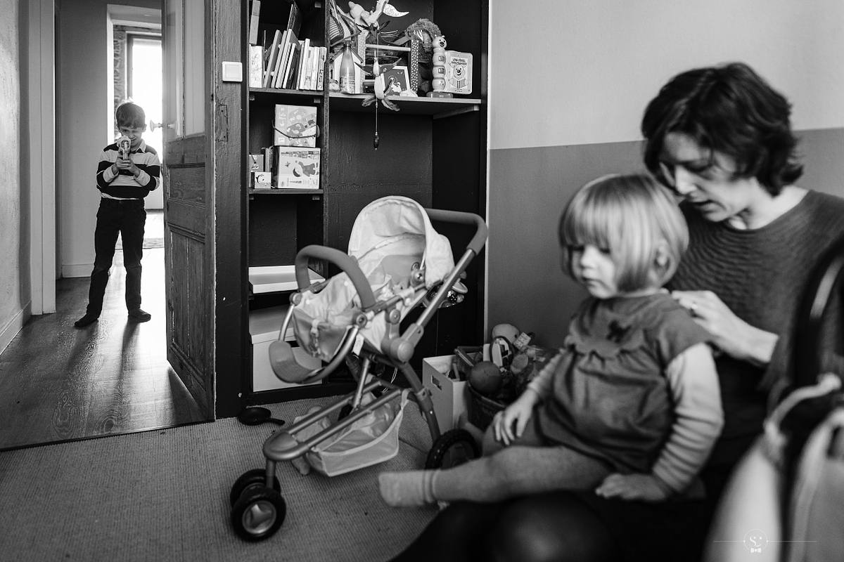 Tarif Photographe Mariage - Sebastien CLAVEL Photographe - Moment intime en famille &agrave; l'int&eacute;rieur avec un enfant jouant dans l'encadrement de la porte et une m&egrave;re soignant son enfant, capturant la vie de famille quotidienne