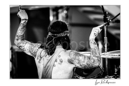 Horizontal black and white image of Mike Portnoy seen from behind at the drum kit with both tattooed arms raised holding drumsticks